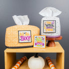 Two Halloween-themed tissue box covers displayed on a wooden shelf against a dark gray wall. One cover is orange and white gingham with a black polka dot trim and a 'Just A Bit BOO' patch, and the other is white with black speckles and a 'This Witch Needs a Tissue' patch.