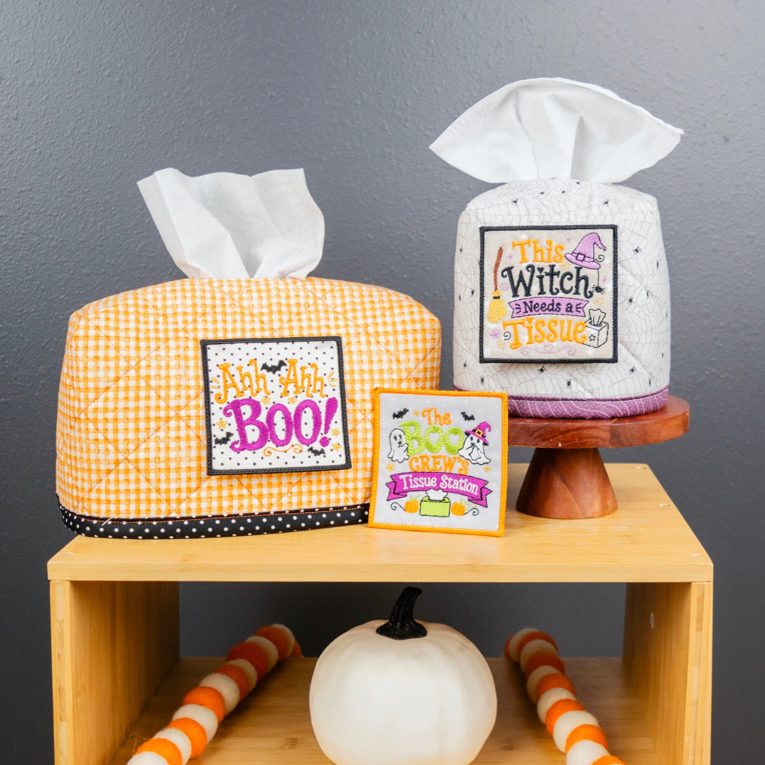Two Halloween-themed tissue box covers displayed on a wooden shelf against a dark gray wall. One cover is orange and white gingham with a black polka dot trim and a 'Just A Bit BOO' patch, and the other is white with black speckles and a 'This Witch Needs a Tissue' patch.
