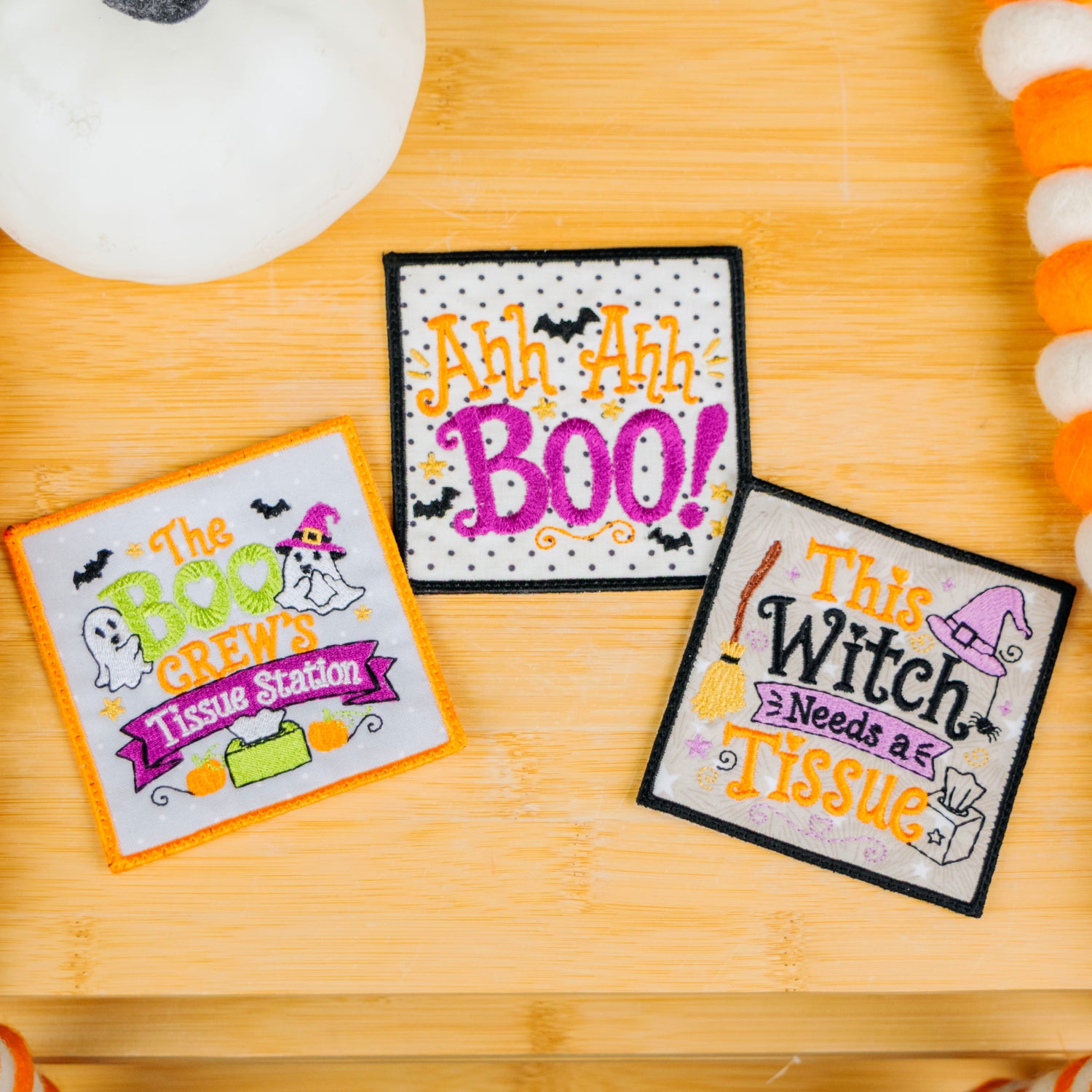 Three Halloween-themed embroidered patches displayed on a wooden surface, surrounded by orange and white striped garland and a white pumpkin decoration.