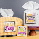 Two Halloween-themed tissue box covers on a wooden table against a dark gray wall. One cover is orange and white gingham with a black polka dot trim and a 'Ah Ah Ah! BOO!' sign, and the other is white with black speckles and a 'This Witch Needs A Tissue' sign.