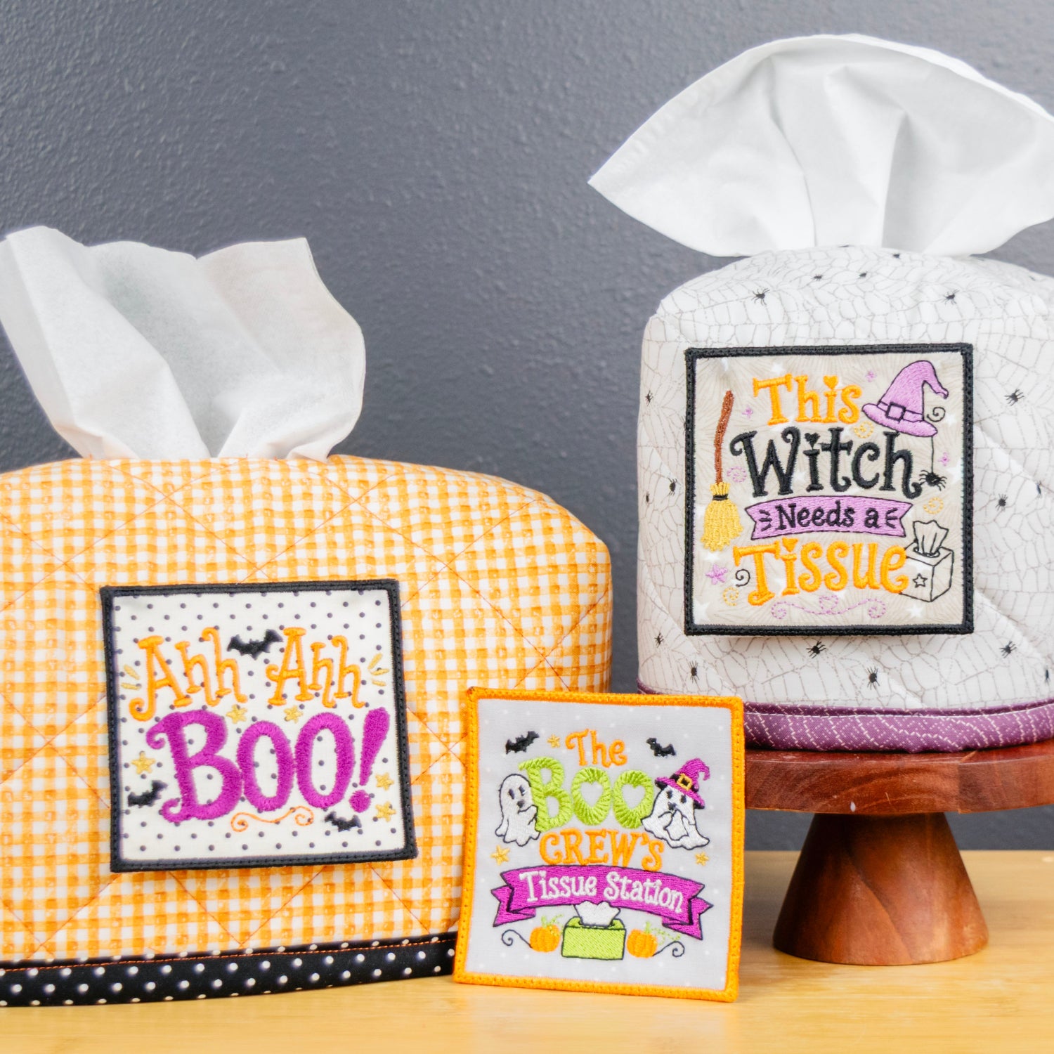 Two Halloween-themed tissue box covers on a wooden table against a dark gray wall. One cover is orange and white gingham with a black polka dot trim and a 'Ah Ah Ah! BOO!' sign, and the other is white with black speckles and a 'This Witch Needs A Tissue' sign.