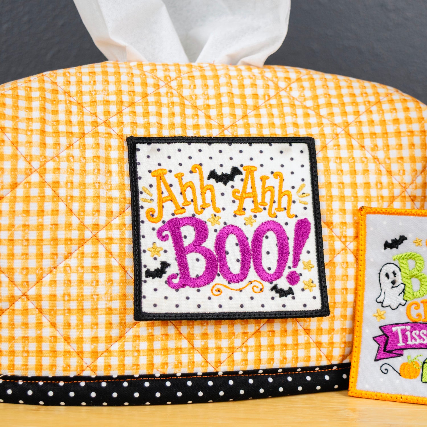 A Halloween-themed tissue box cover in a yellow and white gingham pattern, featuring a black and white polka dot trim and an embroidered patch that reads 'Ah Ah Ah! BOO!'. A smaller embroidered sign next to it reads 'The BOO GREGA Tissue Station'.