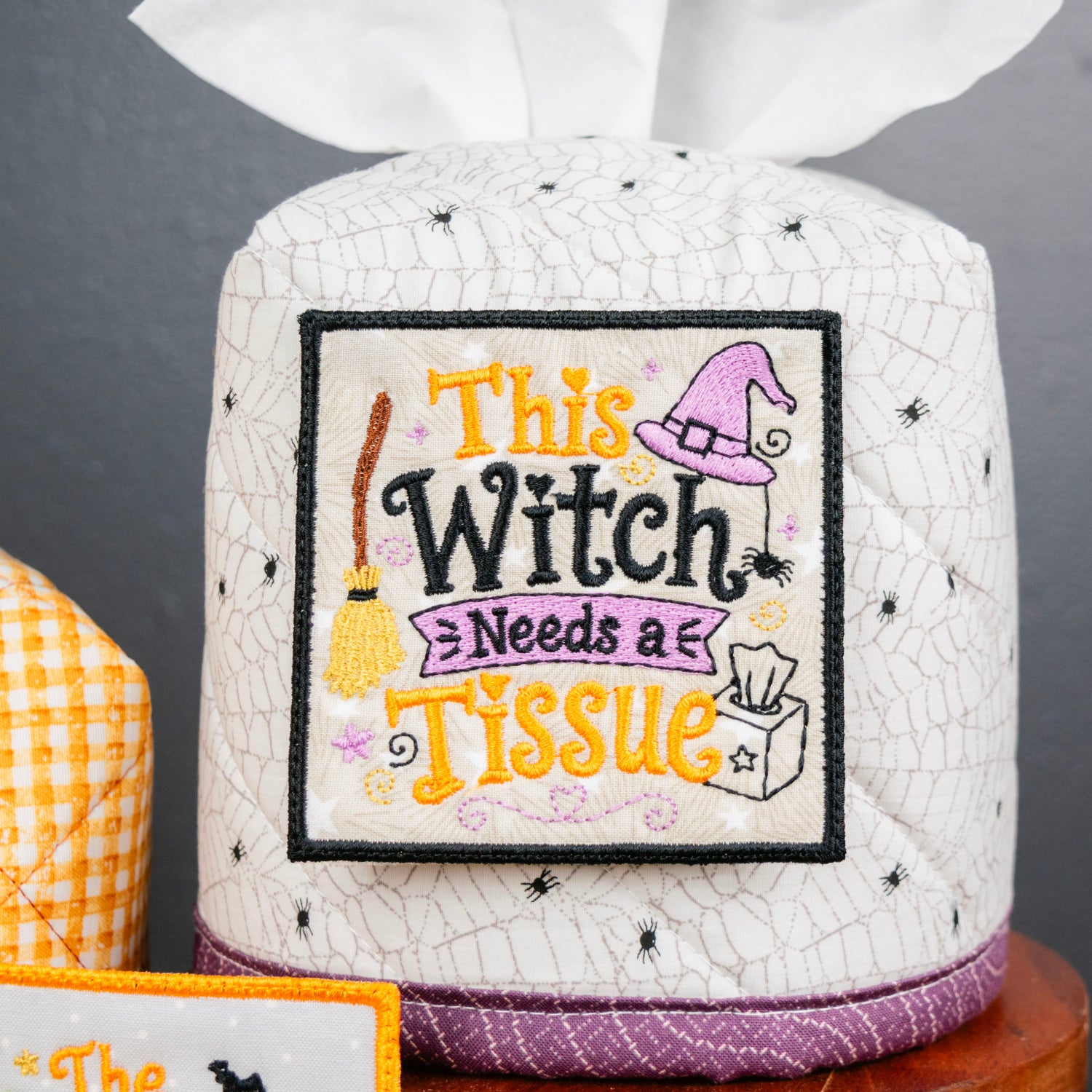 A white tissue box cover with a spiderweb pattern and a central embroidered patch that reads 'This Witch Needs a Issue' with a broom, witch hat, and cat illustrations. A white tissue is partially pulled from the top, and part of another yellow-and-white checkered cover is visible on the left.
