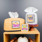 Two Halloween-themed tissue box covers displayed on a wooden shelf against a dark gray wall. One cover is orange and white gingham with a black polka-dot trim and a 'Don't Hype BOO!' patch, and the other is white speckled with a black polka-dot border and a 'This Witch Must Be Obsessed' patch.