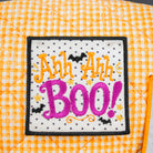A close-up of a square embroidered patch with the phrase “Ah Ah Ah BOO!” in orange and pink lettering on a white background with small black dots and black bats, displayed on an orange-and-white gingham quilted surface.