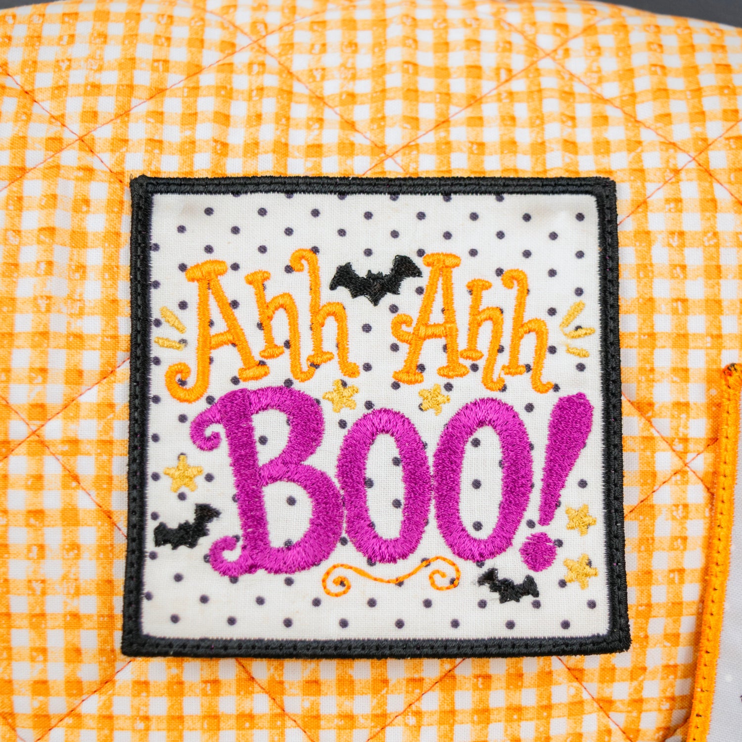 A close-up of a square embroidered patch with the phrase “Ah Ah Ah BOO!” in orange and pink lettering on a white background with small black dots and black bats, displayed on an orange-and-white gingham quilted surface.