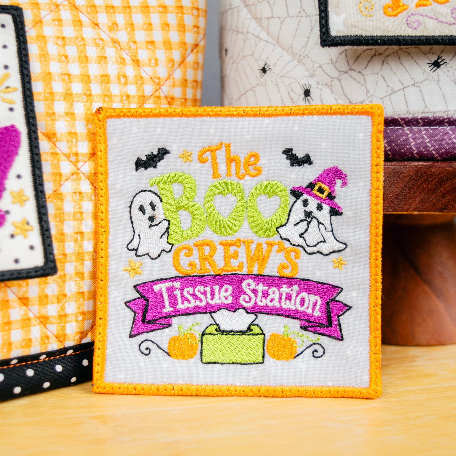 A square embroidered patch with a white background and orange border, featuring the text 'The BOO CREW'S Tissue Station' with cartoon ghosts, bats, pumpkins, and a tissue box. The patch is displayed on a wooden surface next to other Halloween-themed patches.