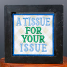 A square black-framed novelty sign with a white center panel reading 'A TISSUE FOR YOUR ISSUE' in blue and green text, displayed on a small wooden stand against a dark gray background.