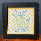 A square black-framed decorative sign with the phrase 'Just ONE of THOSE Days' in blue and green lettering on a yellow patterned background, displayed on a small wooden pedestal against a dark gray backdrop.