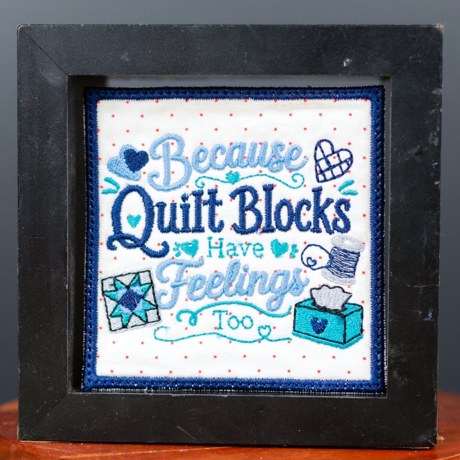 A framed cross-stitch piece with the text 'Because Quilt Blocks Have feelings Too' in blue and white, displayed on a wooden stand against a dark gray background.