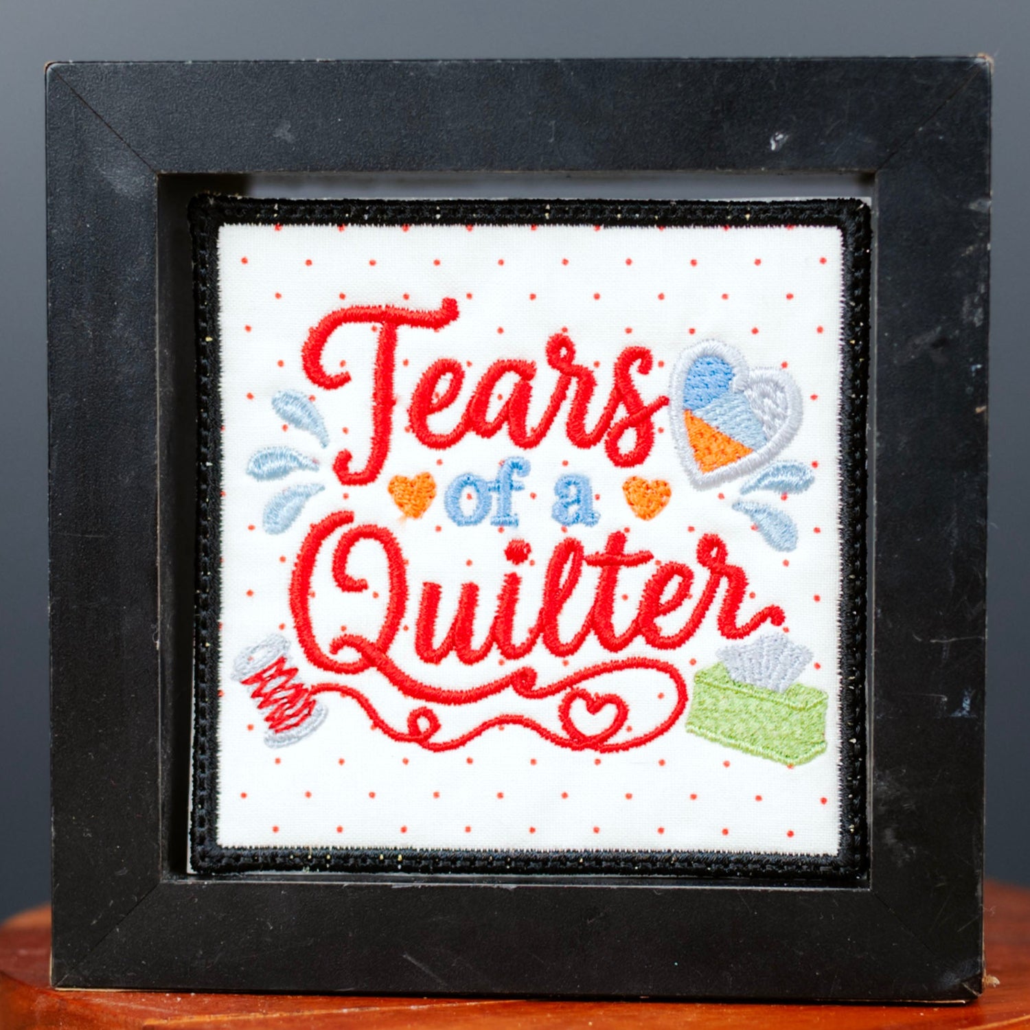 A square black-framed art piece with the words 'Tears of a Quilter' in red and blue lettering, displayed on a small round wooden stand against a dark gray background.