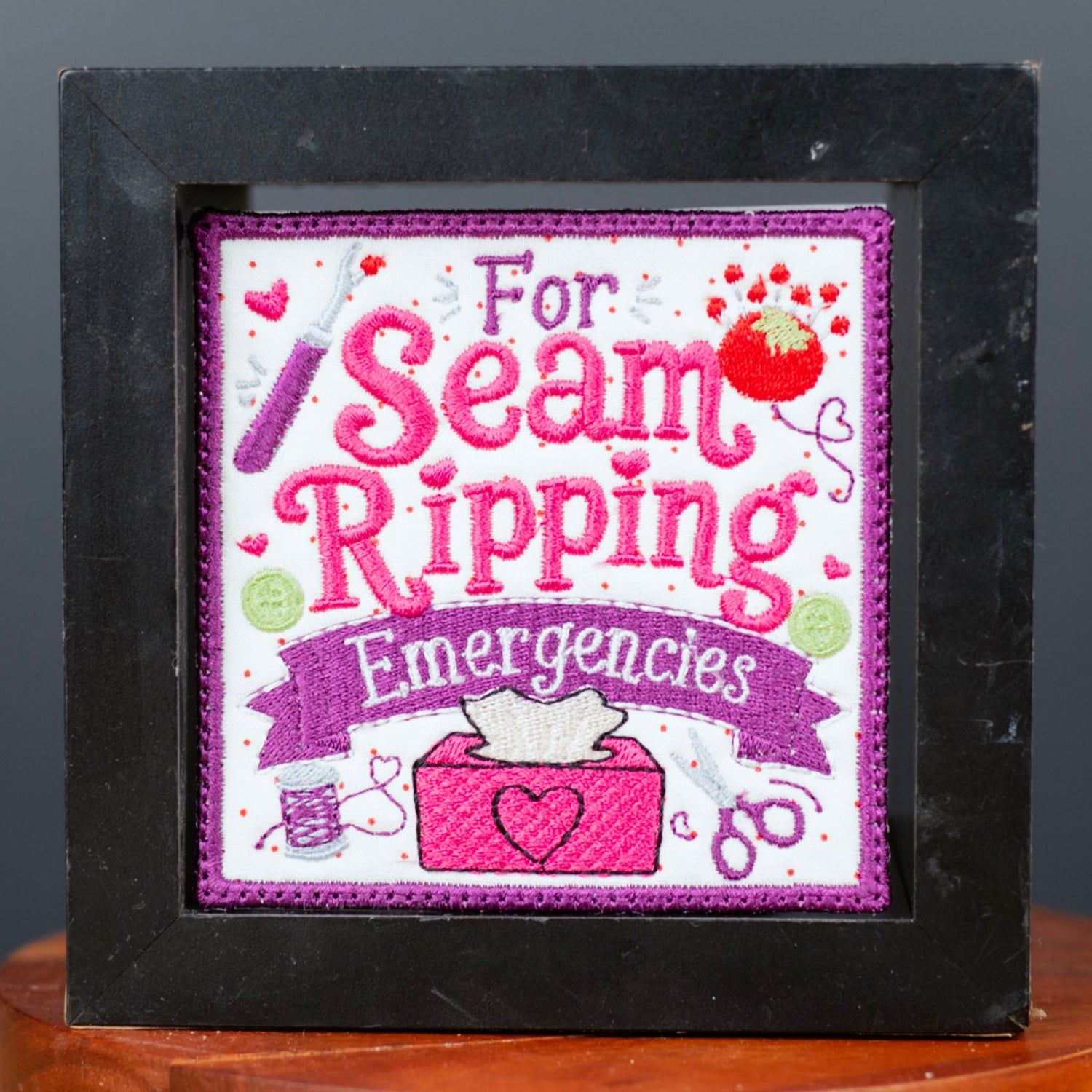 A square black-framed art print or patch with the phrase 'For Seam Ripping Emergencies' in pink and purple lettering, displayed on a small wooden pedestal against a dark gray background.