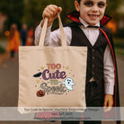 A child in a vampire costume holds up a beige canvas tote bag printed with the phrase 'Too Cute to Spook' in decorative Halloween-themed embroidery, including a ghost, pumpkin, stars, and bats. The background is a blurred outdoor setting with autumn colors.