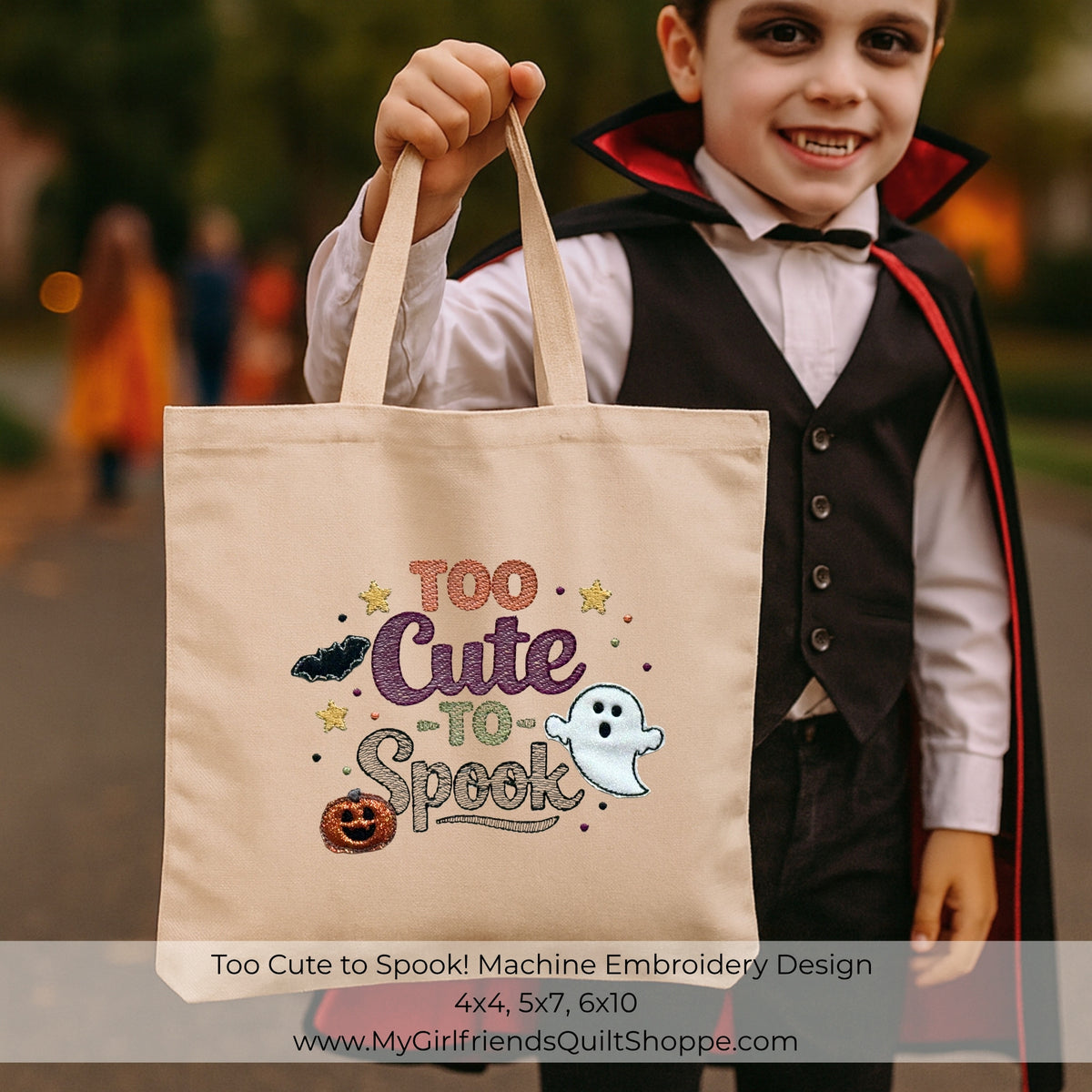 A child in a vampire costume holds up a beige canvas tote bag printed with the phrase 'Too Cute to Spook' in decorative Halloween-themed embroidery, including a ghost, pumpkin, stars, and bats. The background is a blurred outdoor setting with autumn colors.