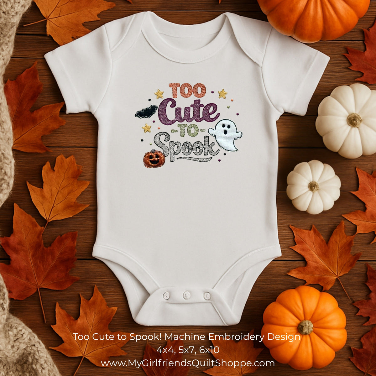 A white short-sleeve baby bodysuit laid flat on a wooden surface, surrounded by orange autumn leaves, small pumpkins, and a beige knit blanket. The bodysuit has a colorful Halloween-themed embroidery design reading “Too Cute to Spook!” with stars, a bat, pumpkin, and ghost motifs.