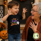 A young boy wearing a black graphic t-shirt that reads 'Too Cute to Spook' in purple, orange, and white, standing next to an older woman in a cozy outdoor setting with pumpkins and fall decorations.