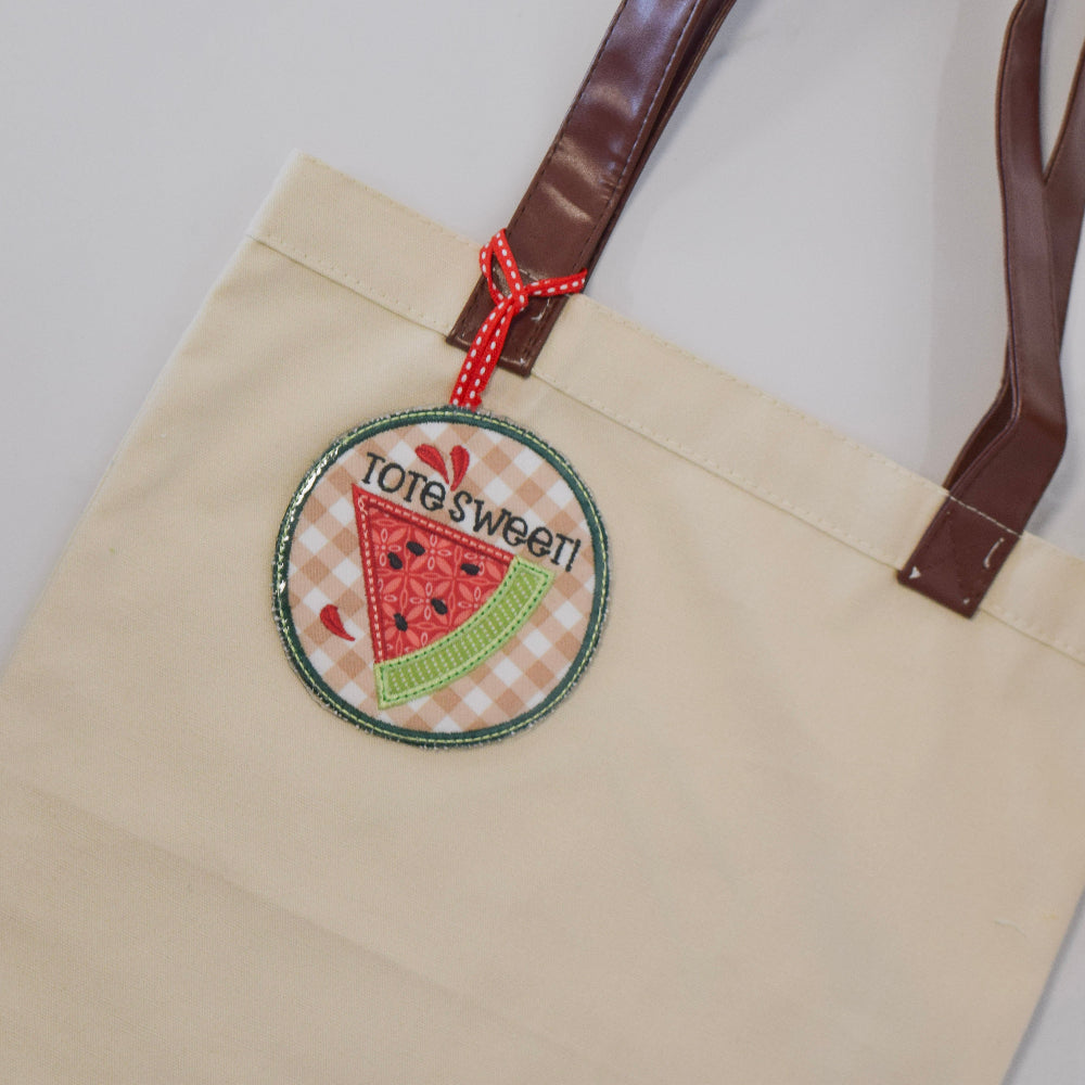 Beige tote bag with a decorative tag featuring a watermelon design on a light background.