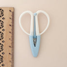 A pair of small scissors with white loop handles and a light blue center section, shown next to a wooden ruler on a plain beige background.