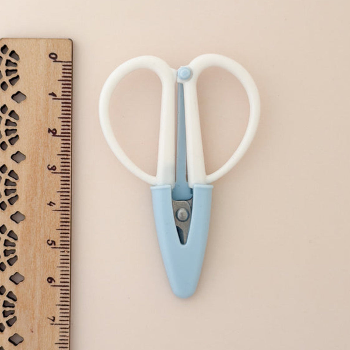A pair of small scissors with white loop handles and a light blue center section, shown next to a wooden ruler on a plain beige background.