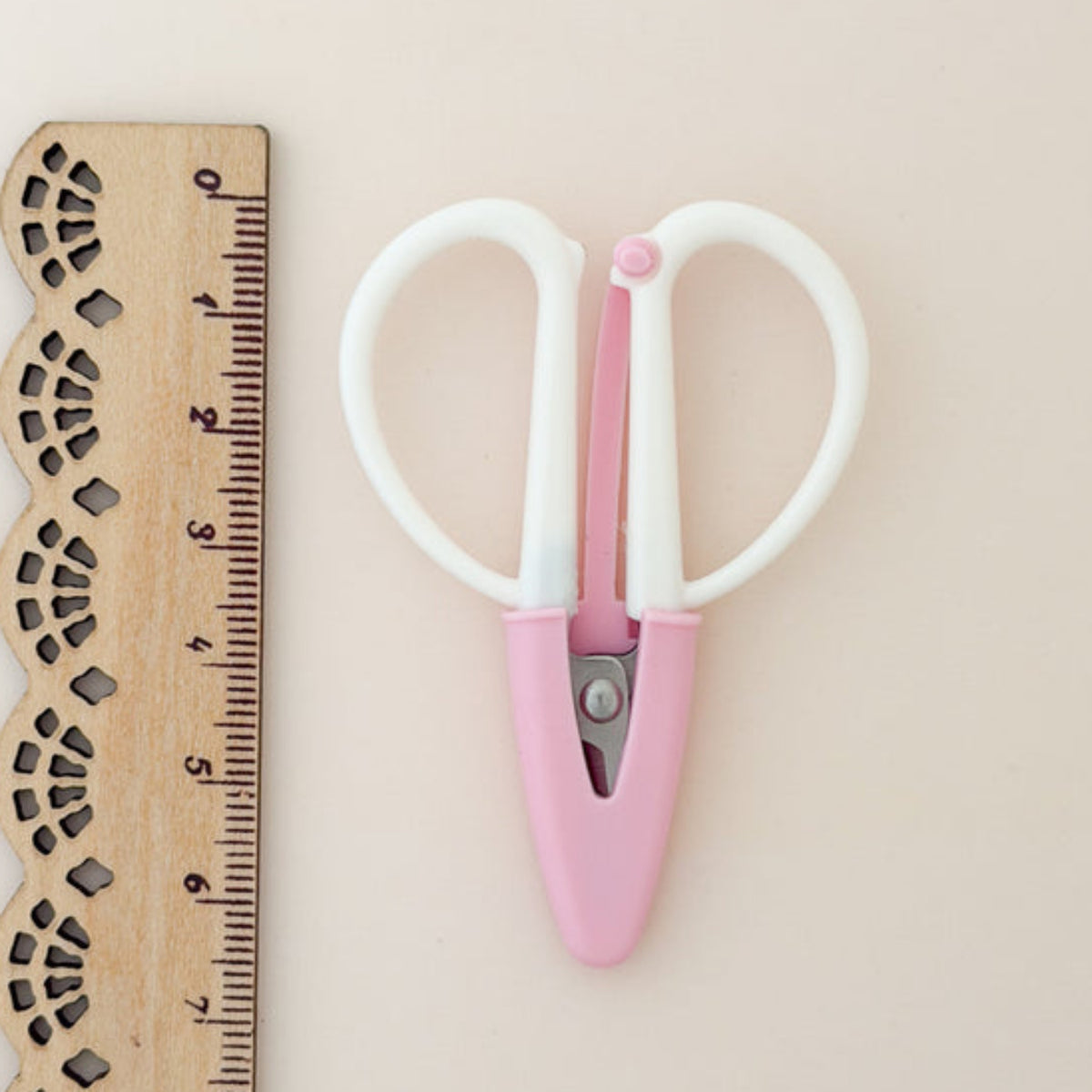 Pink scissors with white handles next to a wooden ruler on a beige background