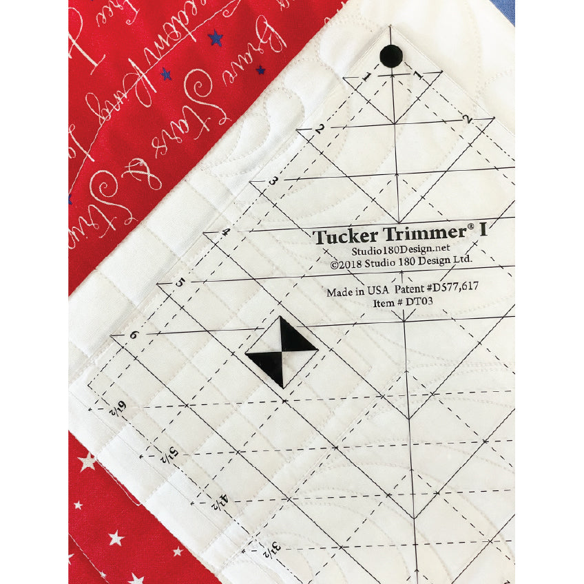 A close-up of a quilting ruler labeled 'Tucker Trimmer II' placed on white quilted fabric, with red patterned fabric visible in the upper left corner.