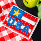 USA-themed patch on a red and white checkered tablecloth with a green mug and red flower in the background.