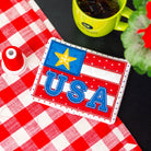 USA-themed patch on a red and white checkered tablecloth with a green mug and flowers.