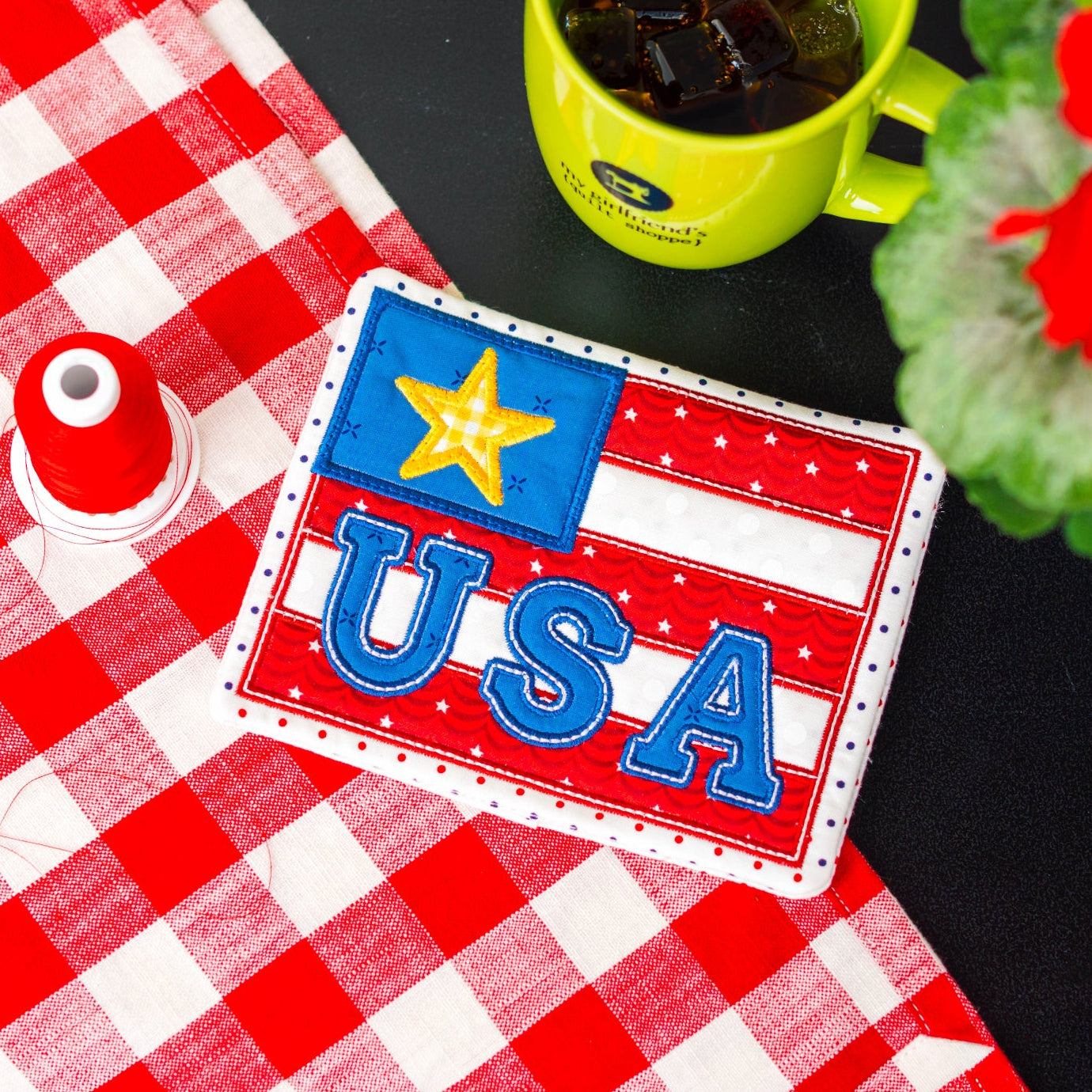 USA-themed patch on a red and white checkered tablecloth with a green mug and flowers.