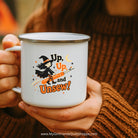 A person in a brown ribbed sweater holds a white enamel mug with a silver rim. The mug features a Halloween graphic of a witch riding a broomstick with the text 'Up, Up, and Unsew!'.