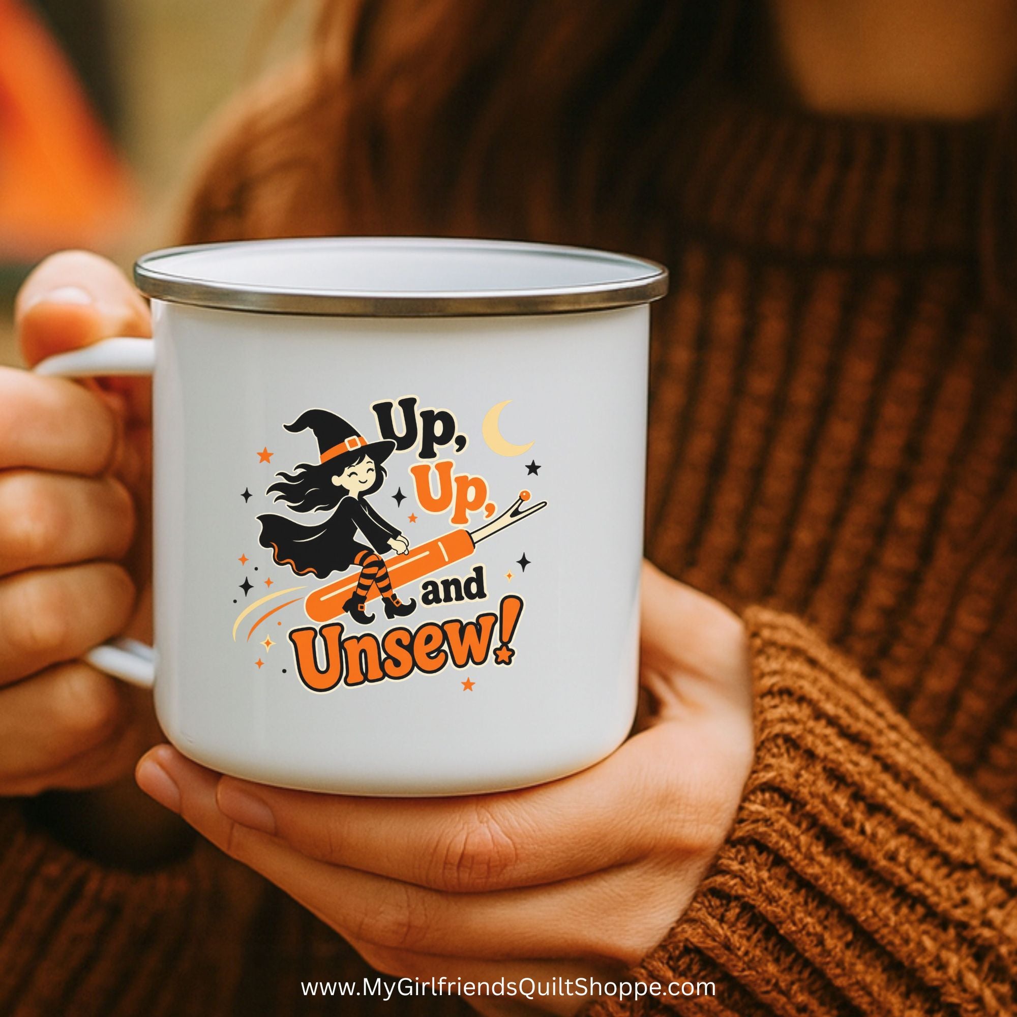 A person in a brown ribbed sweater holds a white enamel mug with a silver rim. The mug features a Halloween graphic of a witch riding a broomstick with the text 'Up, Up, and Unsew!'.