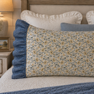 A decorative rectangular pillow with a cream floral pattern and blue ruffled trim, styled on a bed with white and blue pillows in a cozy bedroom setting.