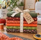 A stack of eight patterned fabric bolts tied with a cream measuring tape ribbon and a kraft paper tag reading 'Fat Quarter Bundle,' displayed on a wooden craft table with sewing-themed props including a BERNINA sewing machine, scissors, a spool of thread, and a cutting mat.