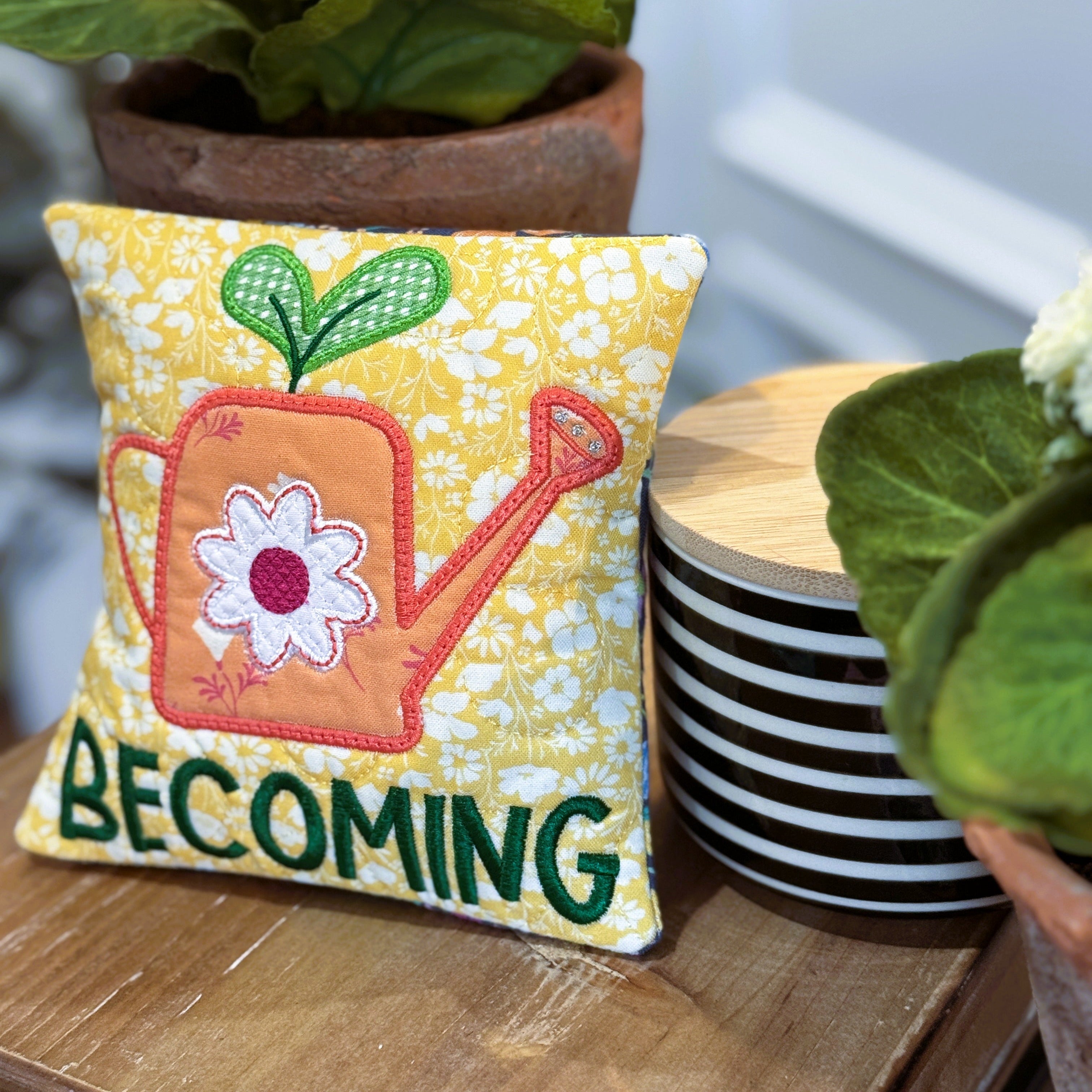 Decorative pillow with a watering can and flower design on a wooden surface with plants in the background.