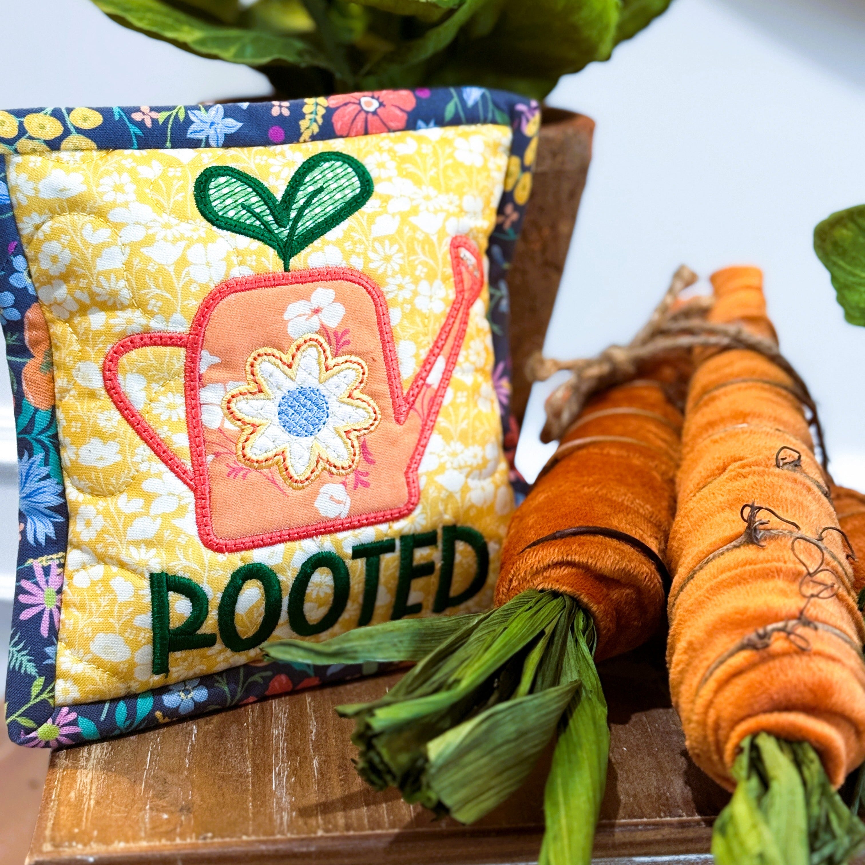 Decorative pillow with 'ROOTED' text and gardening-themed design, surrounded by carrots and flowers.