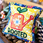 Colorful fabric pillow with 'ROOTED' text and gardening-themed design on a wooden surface.