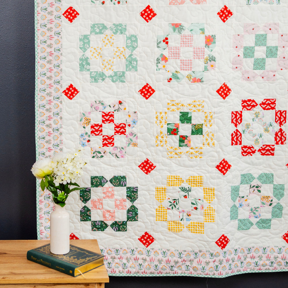 Colorful quilt with geometric patterns on a dark wall, with a wooden table and vase in the foreground.
