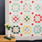 Colorful quilt with geometric patterns on a dark wall, with a wooden table and vase in the foreground.