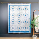 A blue and white SNOWFLAKE patterned quilt hangs from a brass rod against a dark charcoal wall, with a wooden arched hanger and a potted plant to the right.