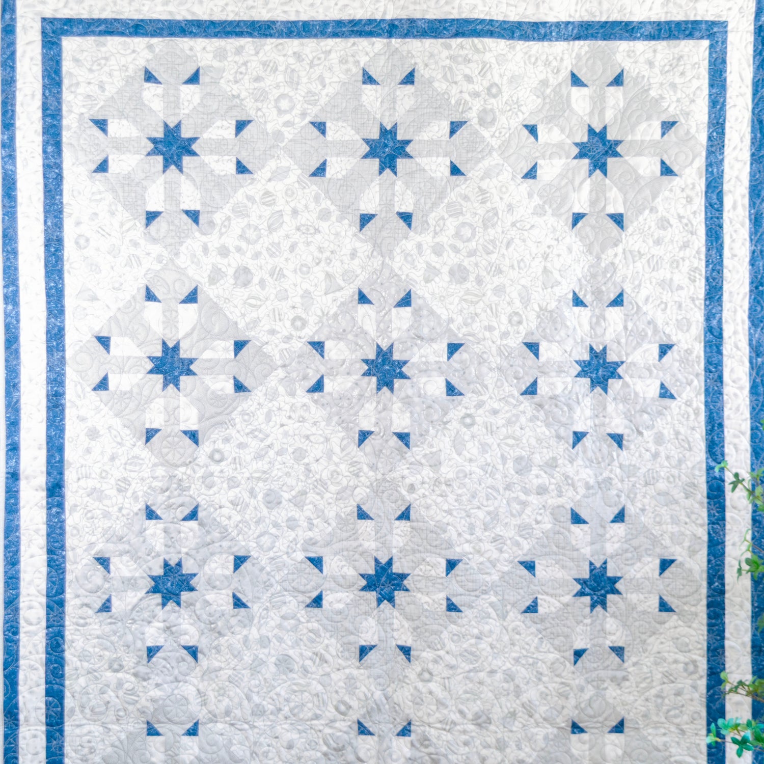 A square quilt hanging flat against a dark gray wall, featuring a white and light gray floral pattern with blue starburst and geometric motifs, bordered by blue and white stripes. A small potted plant and part of a wooden chair are visible at the right edge.