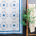A blue and white quilted wall hanging with a geometric star pattern, displayed next to a wooden mirror and a potted green plant against a dark gray wall.