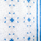 A close-up of a quilted textile featuring a white and light gray floral pattern with blue geometric star and triangle motifs, plus a blue striped border on the right edge.