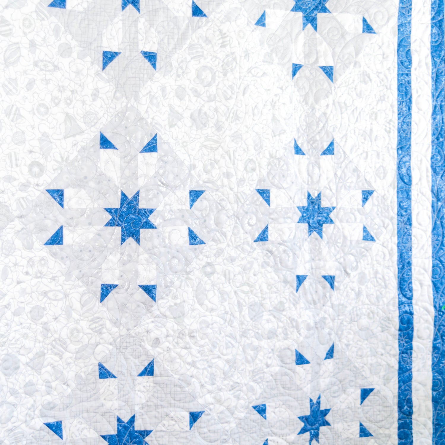 A close-up of a quilted textile featuring a white and light gray floral pattern with blue geometric star and triangle motifs, plus a blue striped border on the right edge.