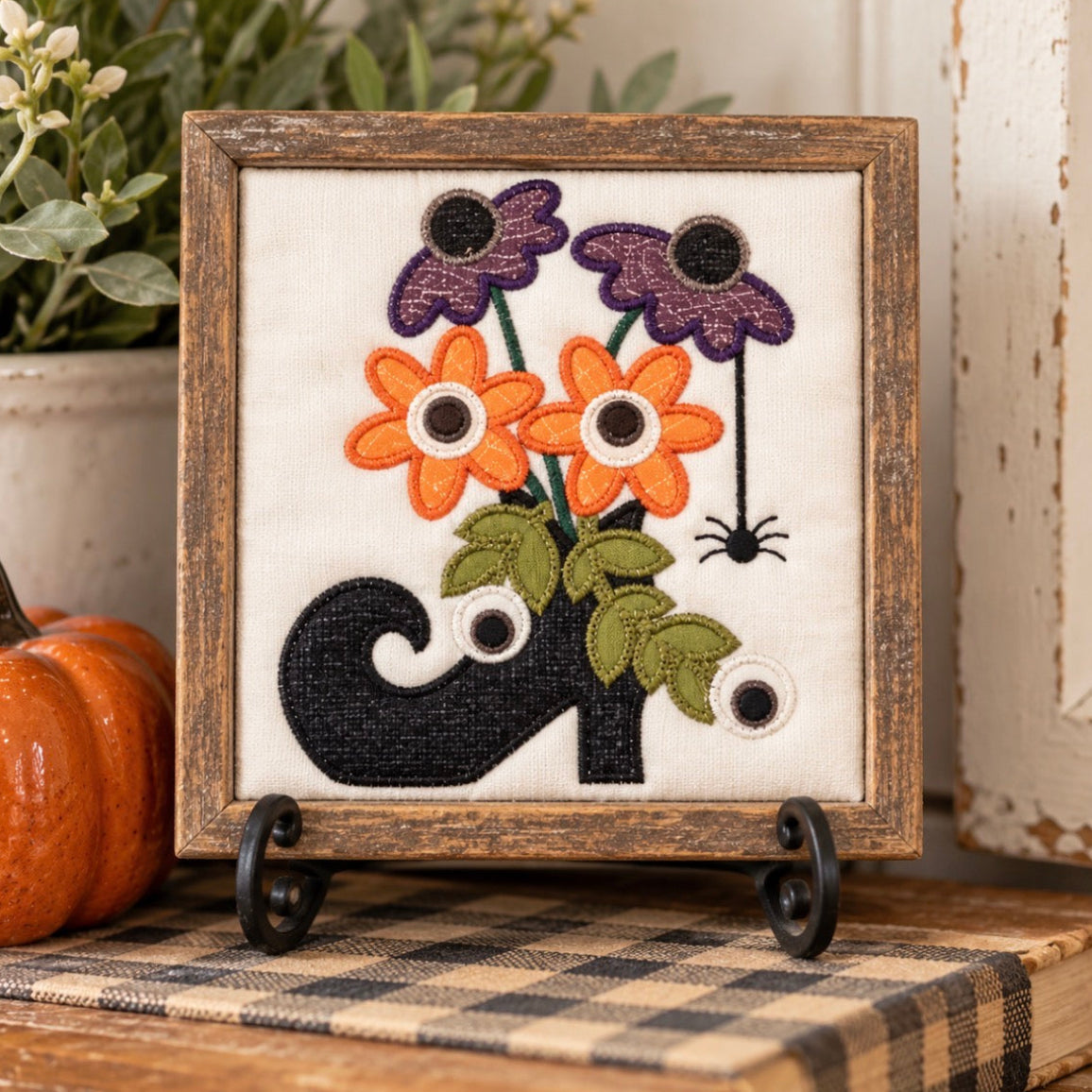 A framed embroidered decorative piece displayed on a small black stand. The artwork shows orange flowers, purple flowers, green leaves, a black cat, and googly eyes, set in a rustic wooden frame on a tabletop with a plaid cloth, a small pumpkin, and plants in the background.