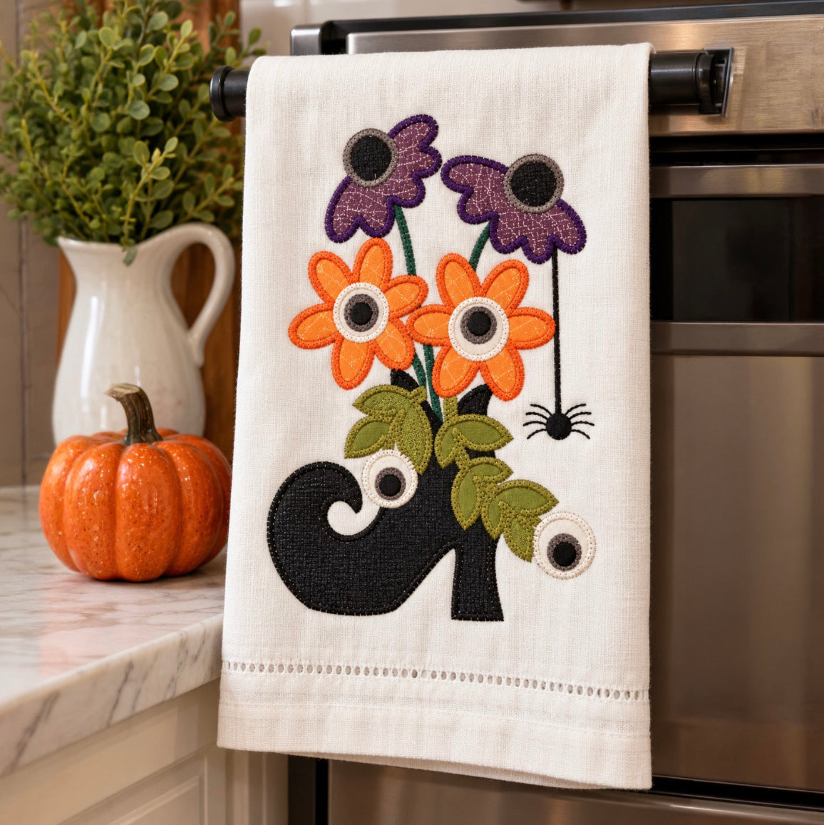 A cream-colored kitchen towel with an embroidered autumn design of orange and purple flowers, green leaves, a black ghost, and a spider, hanging on a black oven handle in a kitchen setting.
