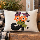 A square decorative throw pillow with a black and white gingham border, featuring an embroidered design of orange and purple flowers, green leaves, and a black boot on a cream linen background. The pillow is styled on a rustic wooden table next to a black and white plaid blanket, with a blurred background of a plant and lantern.