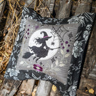 A square decorative pillow with a Halloween-themed embroidered design of a witch riding a broomstick in front of a full moon, surrounded by spiders, cobwebs, and purple flowers. The pillow features a black and gray floral border and is displayed on a rustic wooden surface.