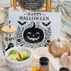 A styled Halloween tabletop scene with a white sign reading “Happy Halloween” and a large black jack-o'-lantern, surrounded by pumpkins, a bowl of lime slices, candy, sprinkles, and decorative textiles. A framed sign in the background lists craft names.
