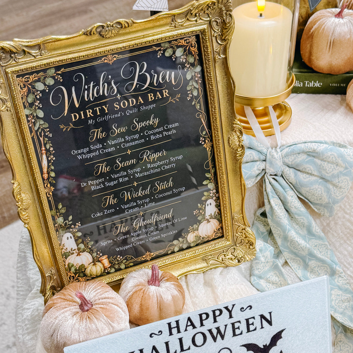 Halloween-themed tabletop decor featuring a gold-framed 'Witch's Brew Dirty Soda Bar' sign, a light blue 'Happy Halloween' card with a jack-o'-lantern, pumpkins, a lit candle, and a tied fabric bow.