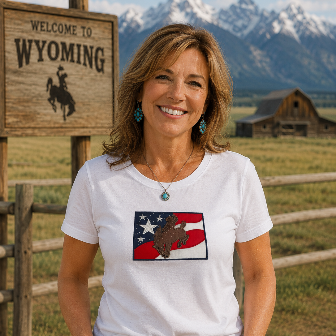 Woman wearing a white t-shirt with a graphic design, standing in front of a 'Welcome to Wyoming' sign and mountain landscape.