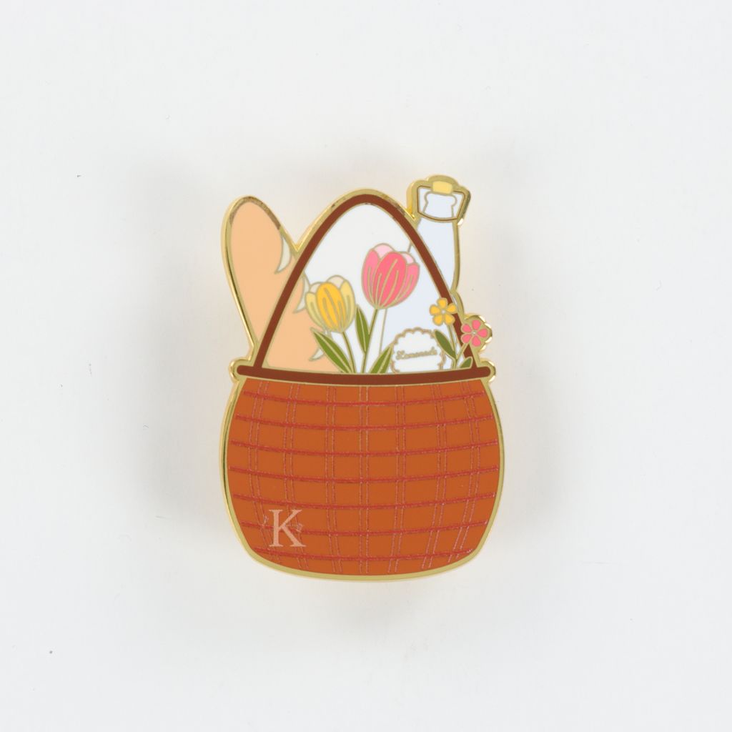 Enamel pin of a picnic basket with bread, flowers, and a bottle on a white background